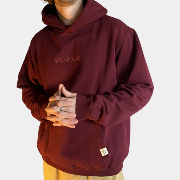 A person wearing a burgundy Shakahs hoodie with the brand's logo embroidered on the front and the sleeve.